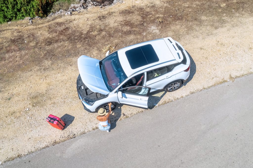 A broken down car on the side of the road with the bonnet open. A woman with her suitcase is sat checking the car.