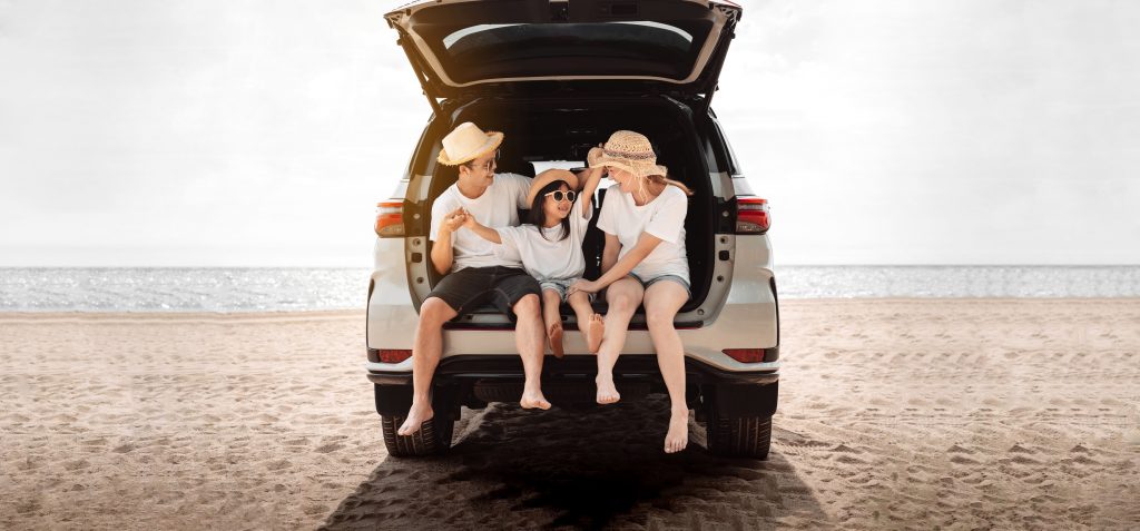 A family of three going on a summer holiday. They are sat in the boot of a car on a beach.
