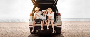 A family of three going on a summer holiday. They are sat in the boot of a car on a beach.